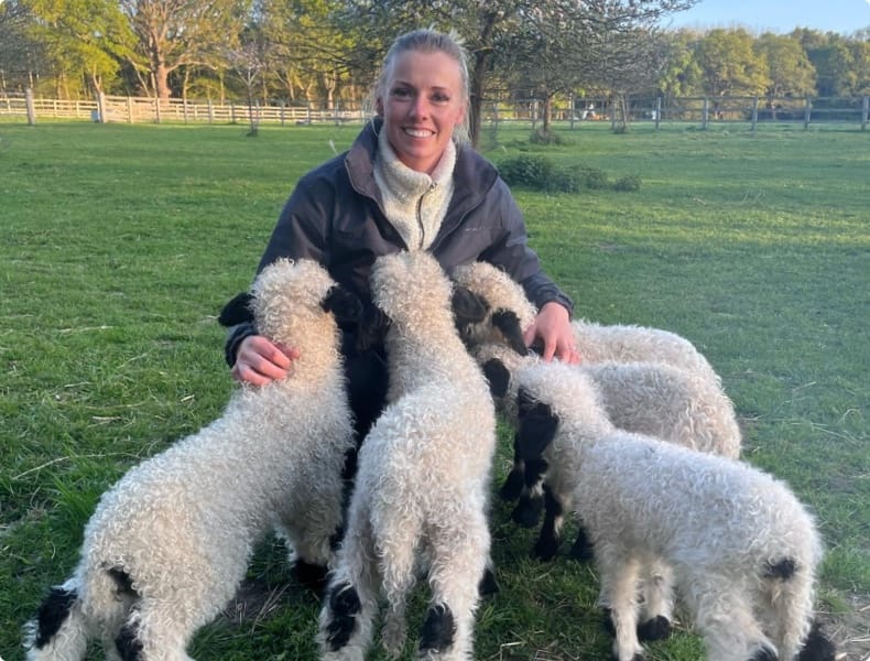 Rebecca smiles at the camera while lambs nuzzle at her jacket